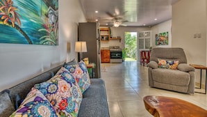 A living room with a sofa, armchair, wooden coffee table, and a kitchen area with a stove and cabinets.