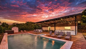 A pool area with lounge chairs and a covered seating area, surrounded by trees.
