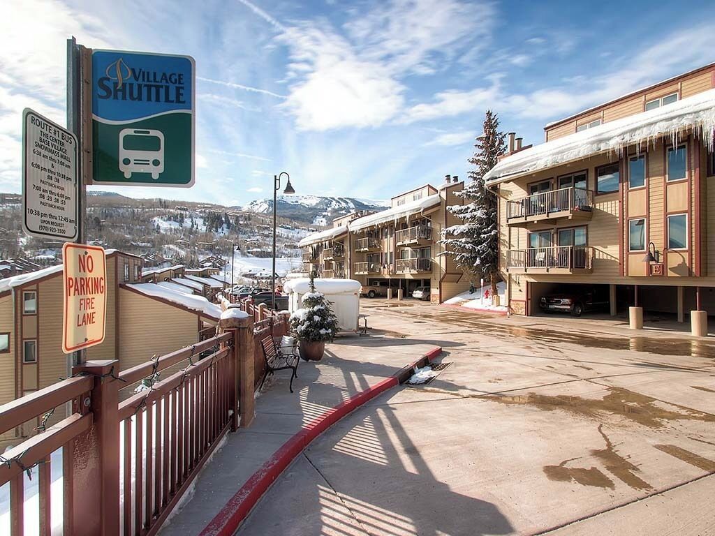 Señal de servicio de transporte en el pueblo, señal de prohibido estacionar en zona de acceso para vehículos de emergencia, y vista de un edificio cubierto de nieve con balcones.