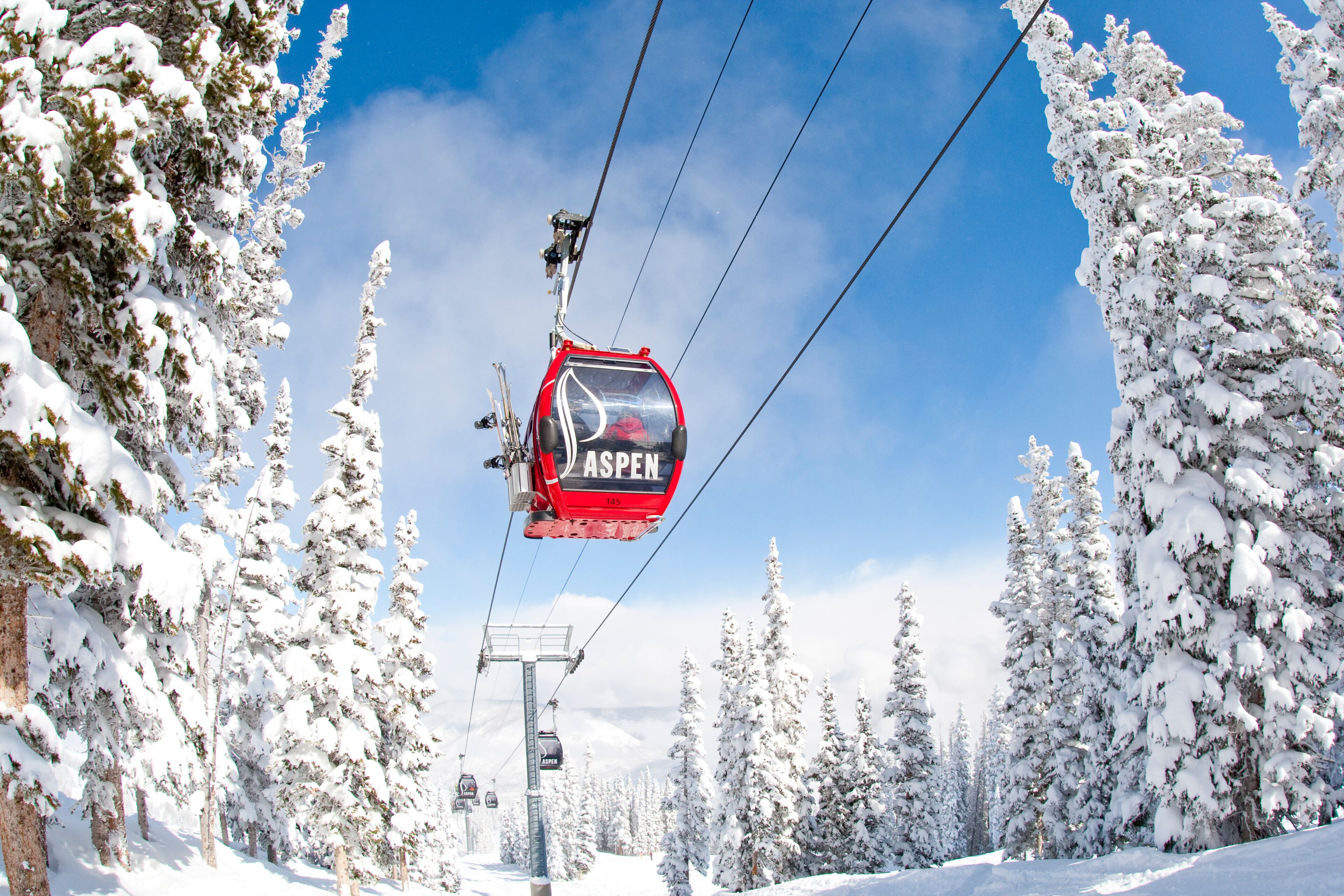 Eine Seilbahn mit einer roten Kabine mit dem Aufdruck "ASPEN", die an Seilen hängt, umgeben von schneebedeckten Bäumen.