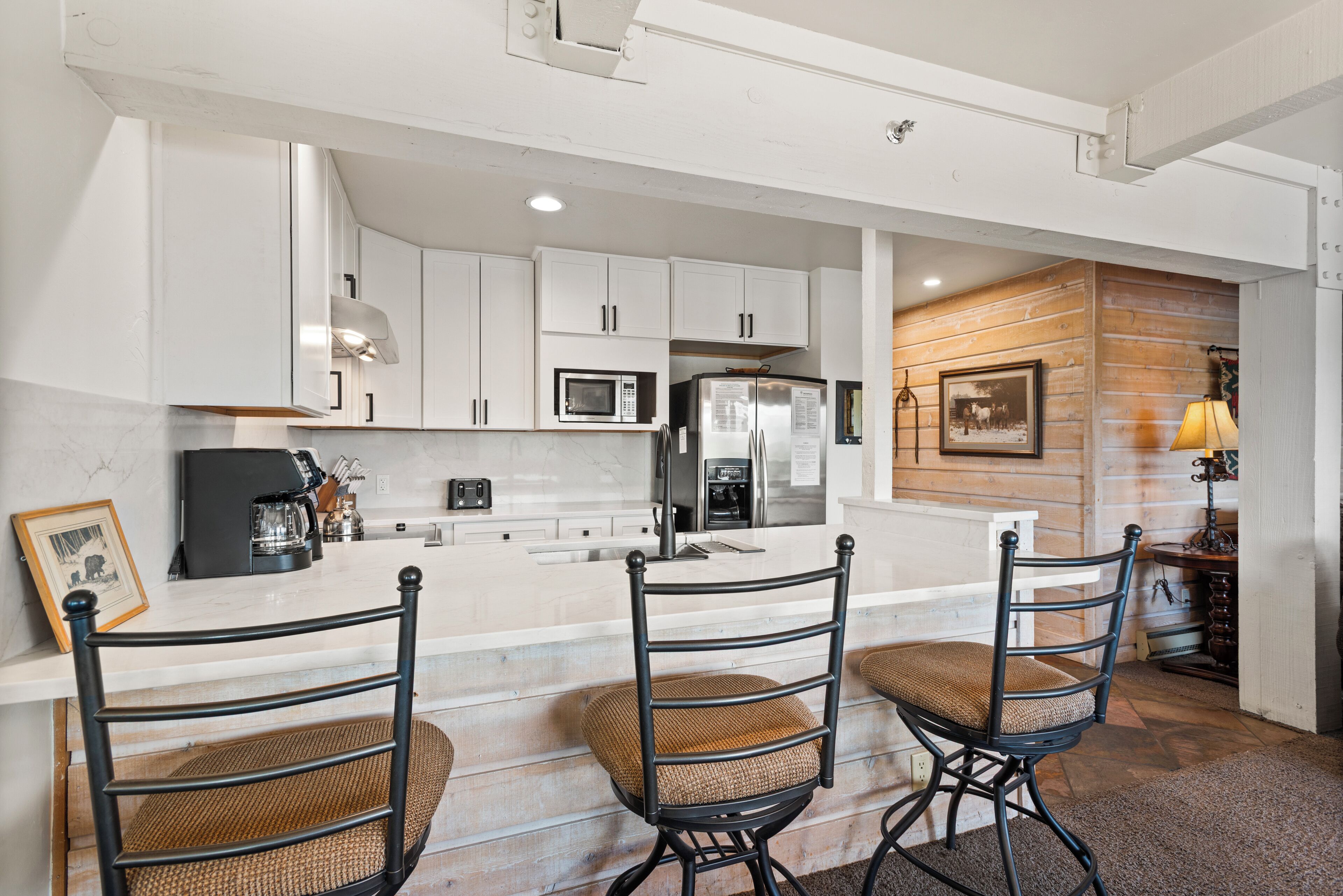 A kitchen with white cabinets, a microwave, a refrigerator, and a coffee maker. There are three wicker chairs with cushions.