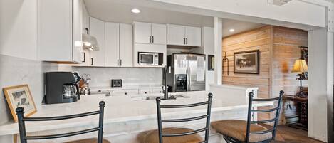 A kitchen with white cabinets, a microwave, a refrigerator, and a coffee maker. There are three wicker chairs with cushions.