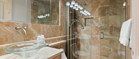 Ein Badezimmer mit einem Waschbecken aus Glas, einer Toilette, einer Dusche mit Glastür und einem beleuchteten Spiegel.