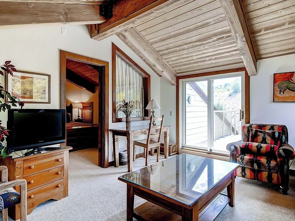 Un vaste salon doté d’un plafond en bois, d’une table basse en verre, d’un téléviseur, d’un coin repas avec une table et des chaises, et d’une grande fenêtre offrant une vue sur l’extérieur.