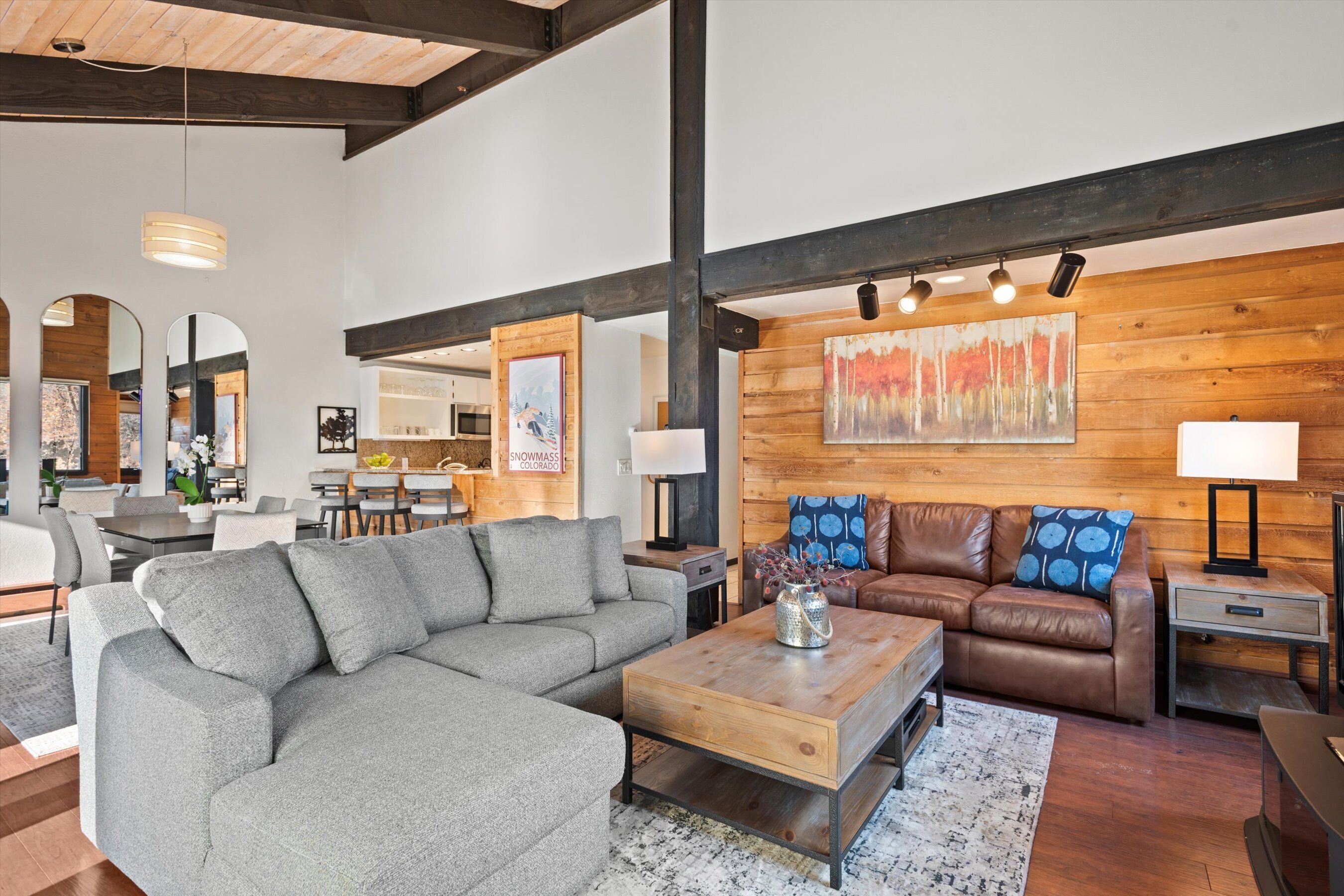 A spacious living area with a large gray sectional sofa, a brown leather sofa, and a wooden coffee table. There is a painting on the wall and a kitchen area in the background.