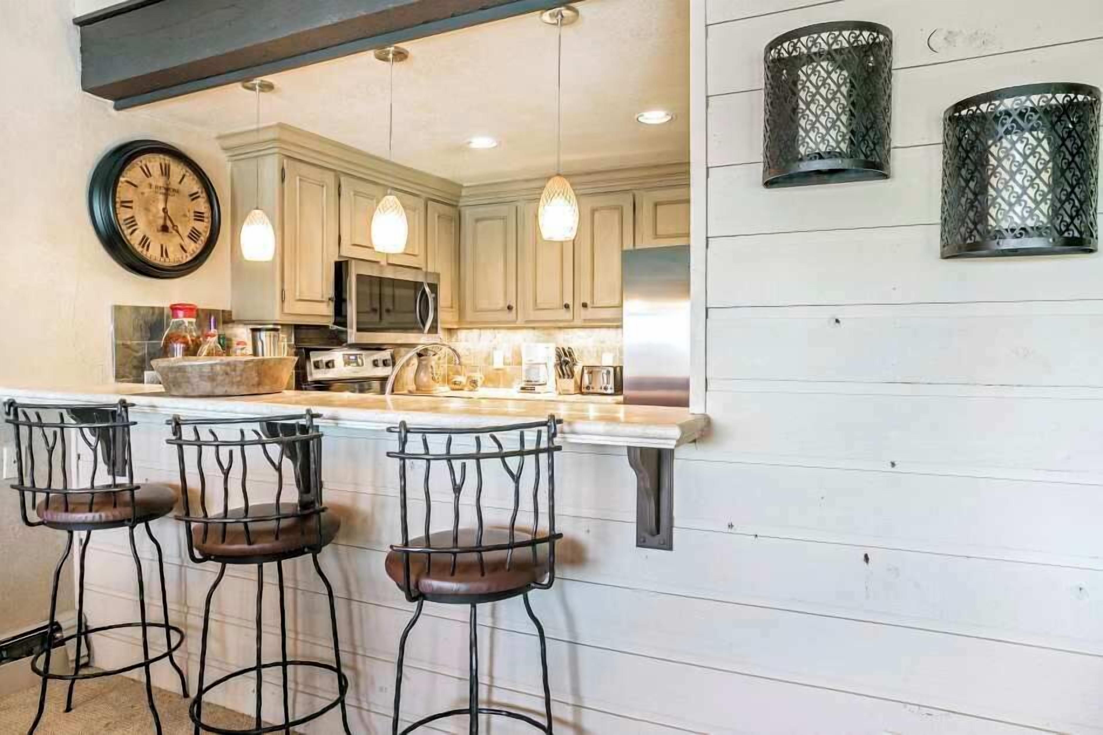 A kitchen with white cabinets, a microwave, a stove, and a sink. There are three bar stools with metal frames and leather seats. A clock is mounted on the wall above the counter.