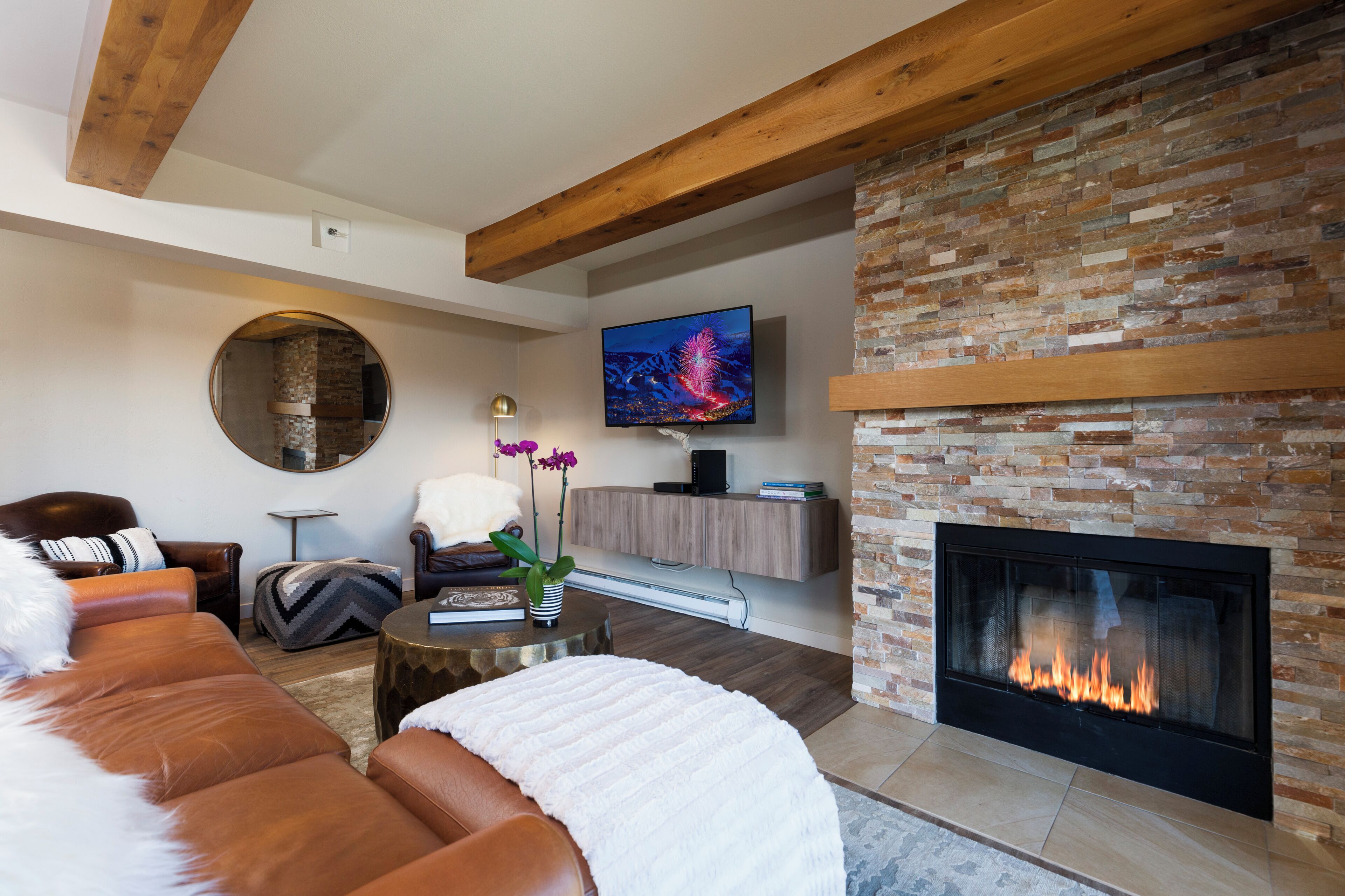 A modern living room with a fireplace, leather sofa, round mirror, and a flat-screen TV.