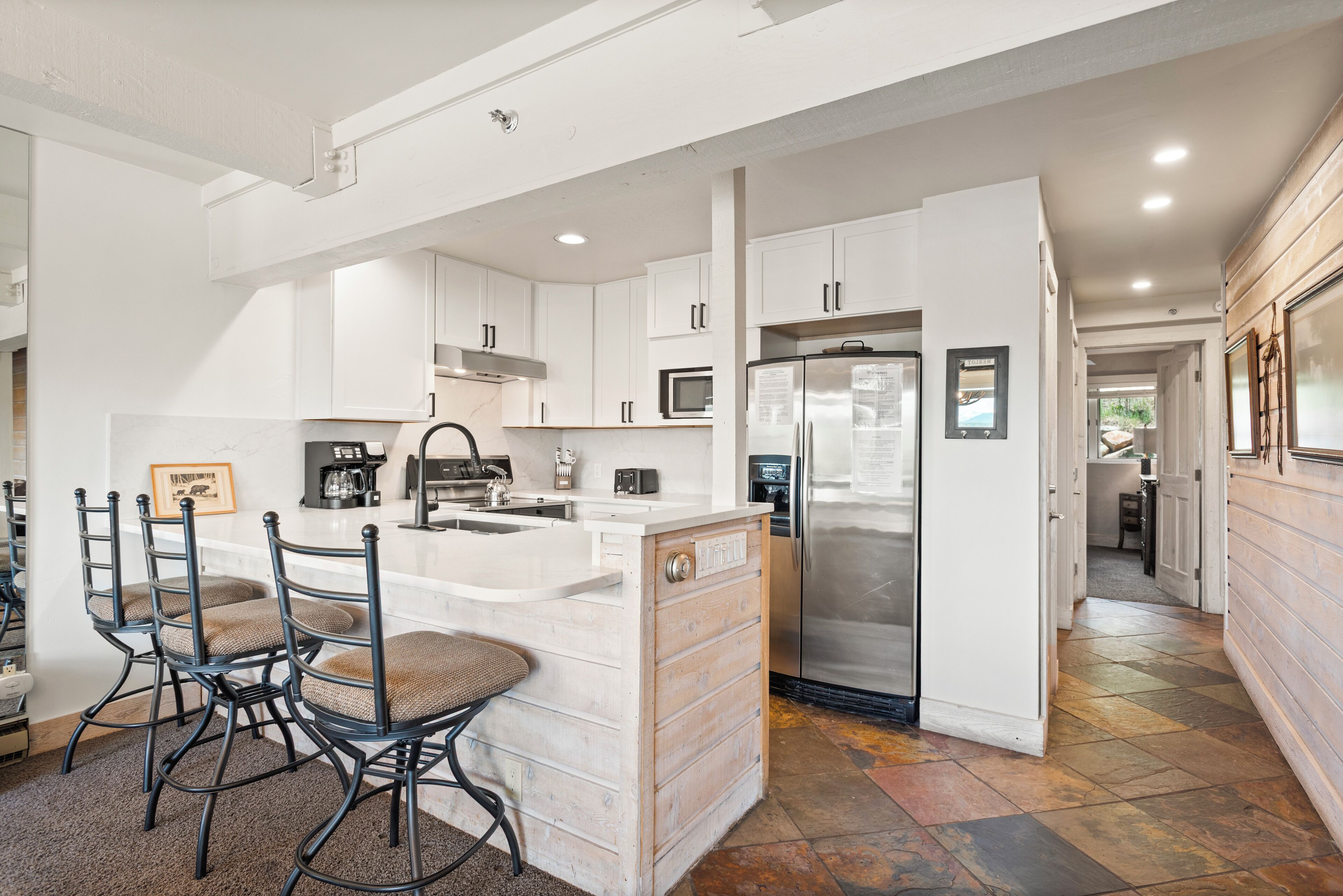 A modern kitchen with white cabinetry, a central island with bar stools, a stainless steel refrigerator, and a microwave.