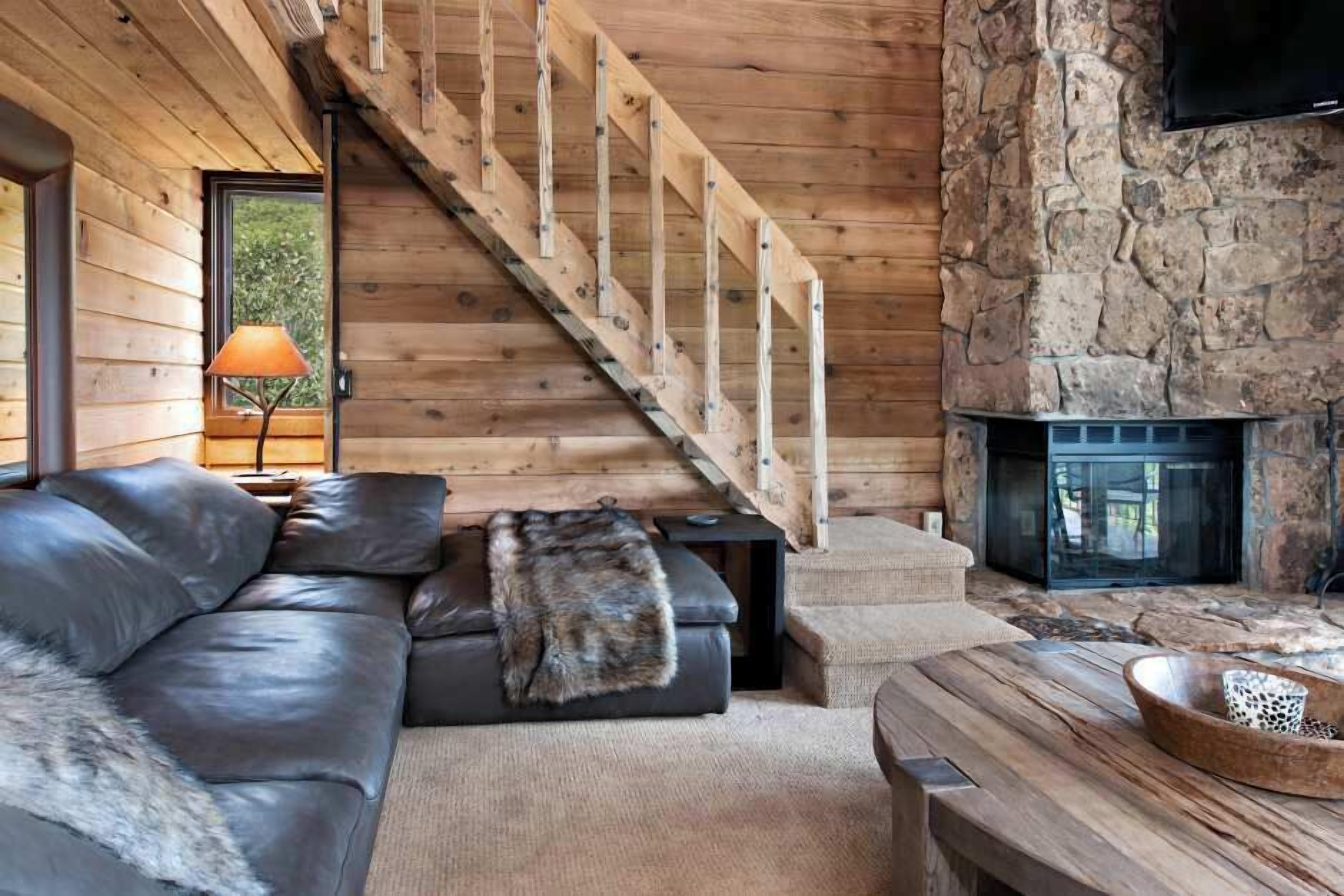 A log cabin interior with a wooden staircase, a stone fireplace, a leather sofa, and a rustic dining table.