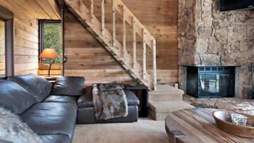 A log cabin interior with a wooden staircase, a stone fireplace, a leather sofa, and a rustic dining table.