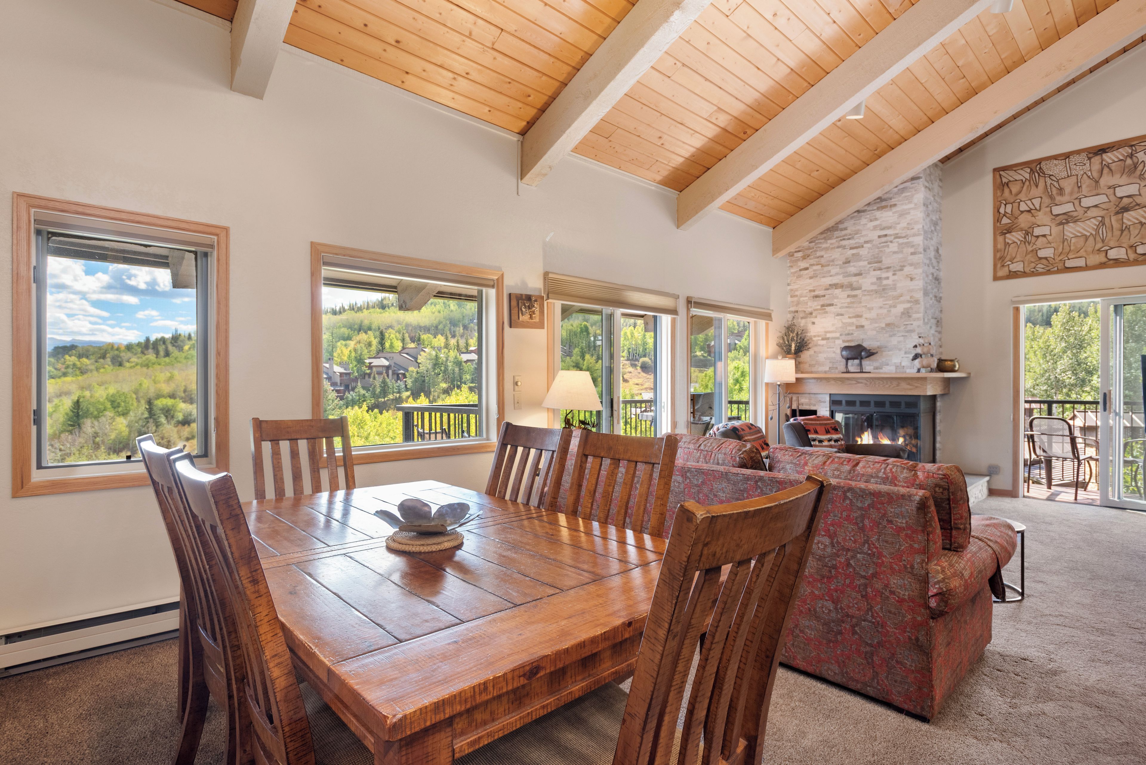 A spacious dining area with a large wooden table and chairs, large windows offering a view of the outdoors, and a stone fireplace.