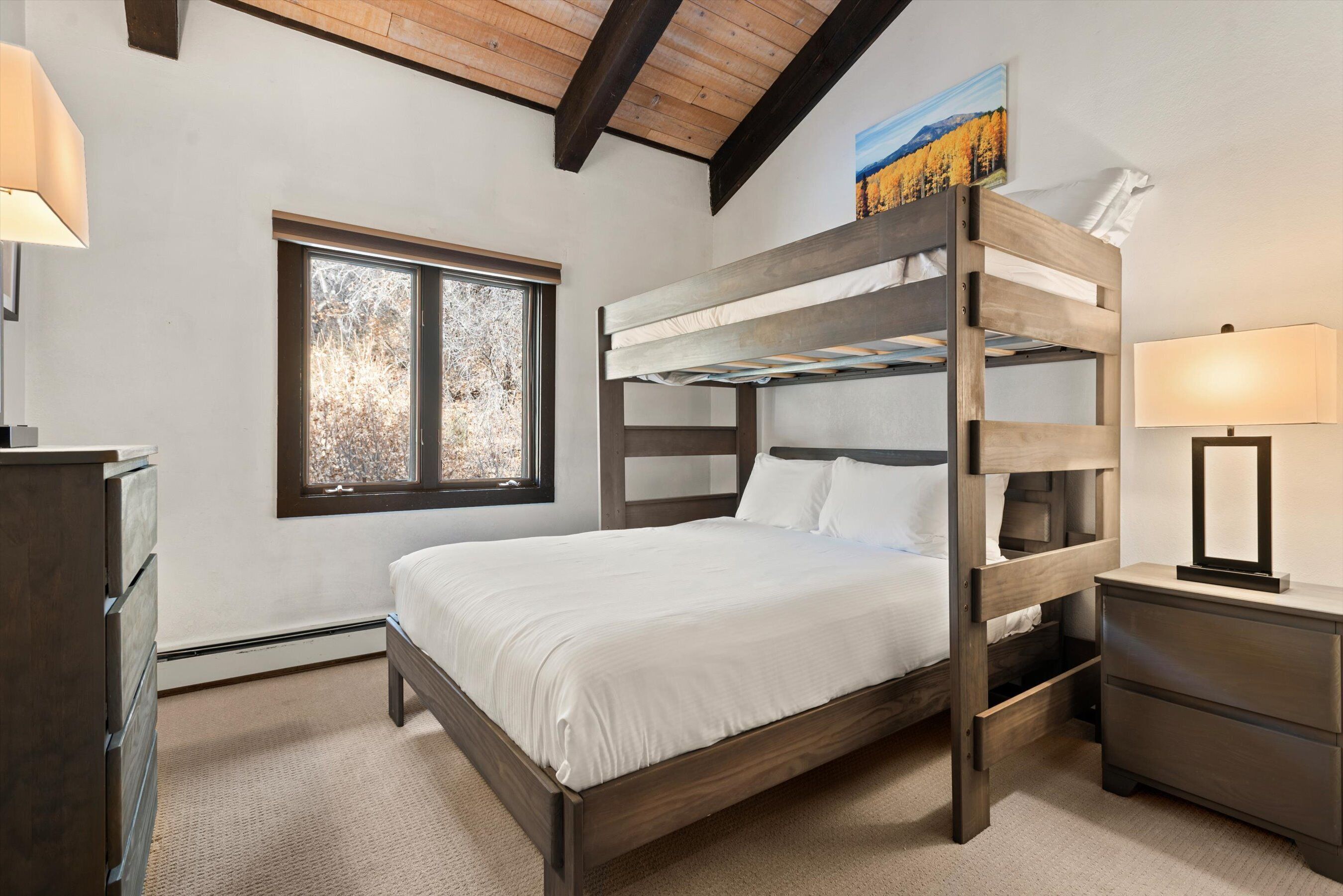 A bedroom with a bunk bed, a window with a view of trees, a dresser, and a lamp.