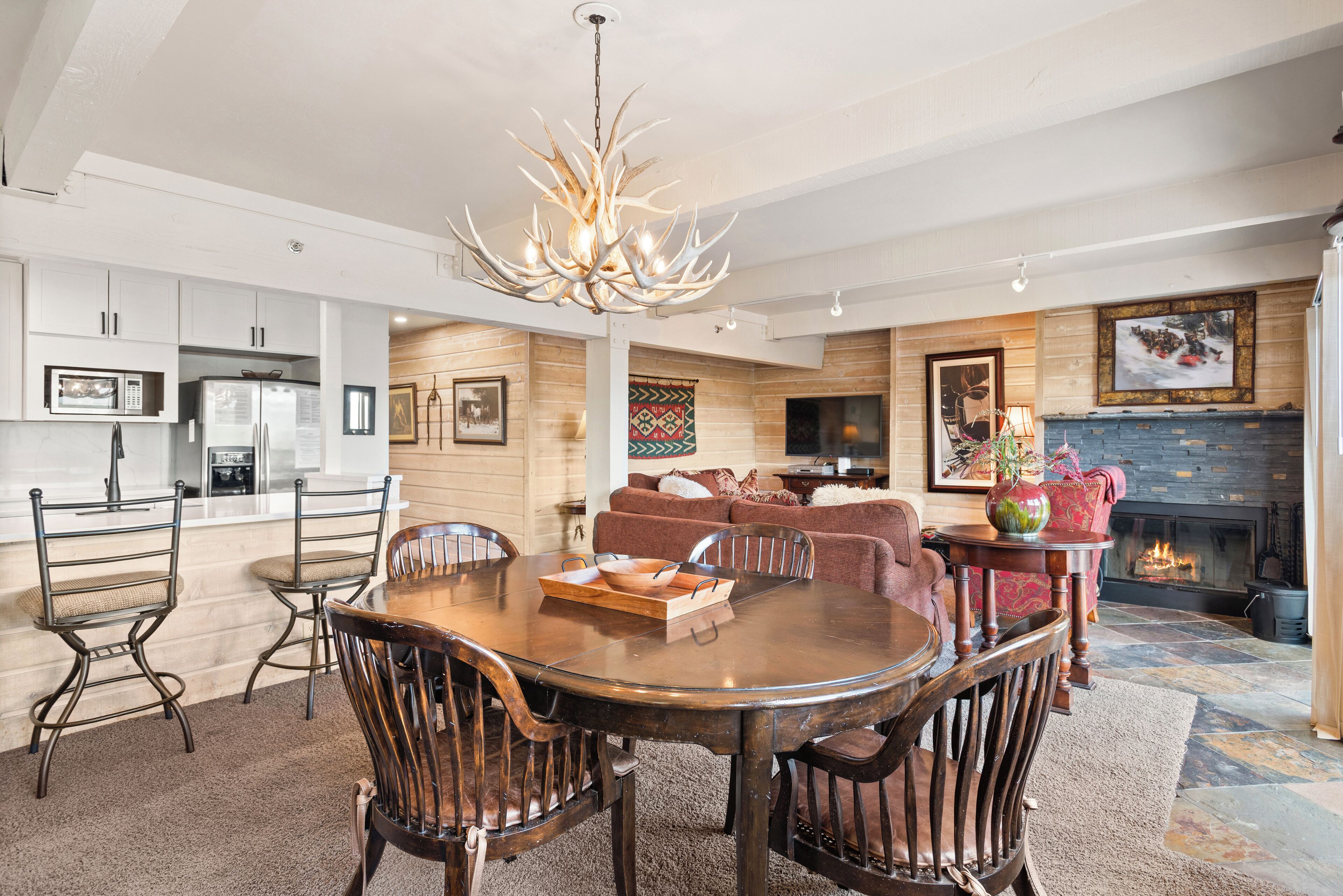 A modern kitchen with white cabinets, a dining area with a wooden table and chairs, a fireplace, and a living room with a sofa and a TV.