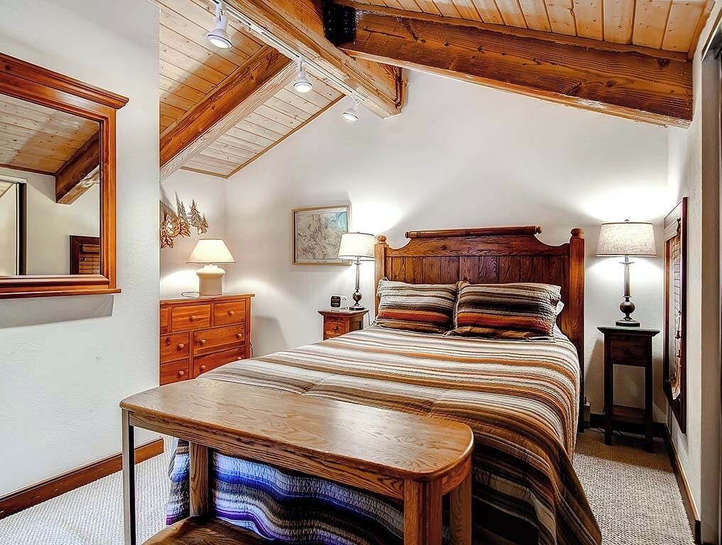 Un dormitorio con techo de madera, una cama con ropa de cama a rayas, una mesita de noche con lámpara y un aparador.