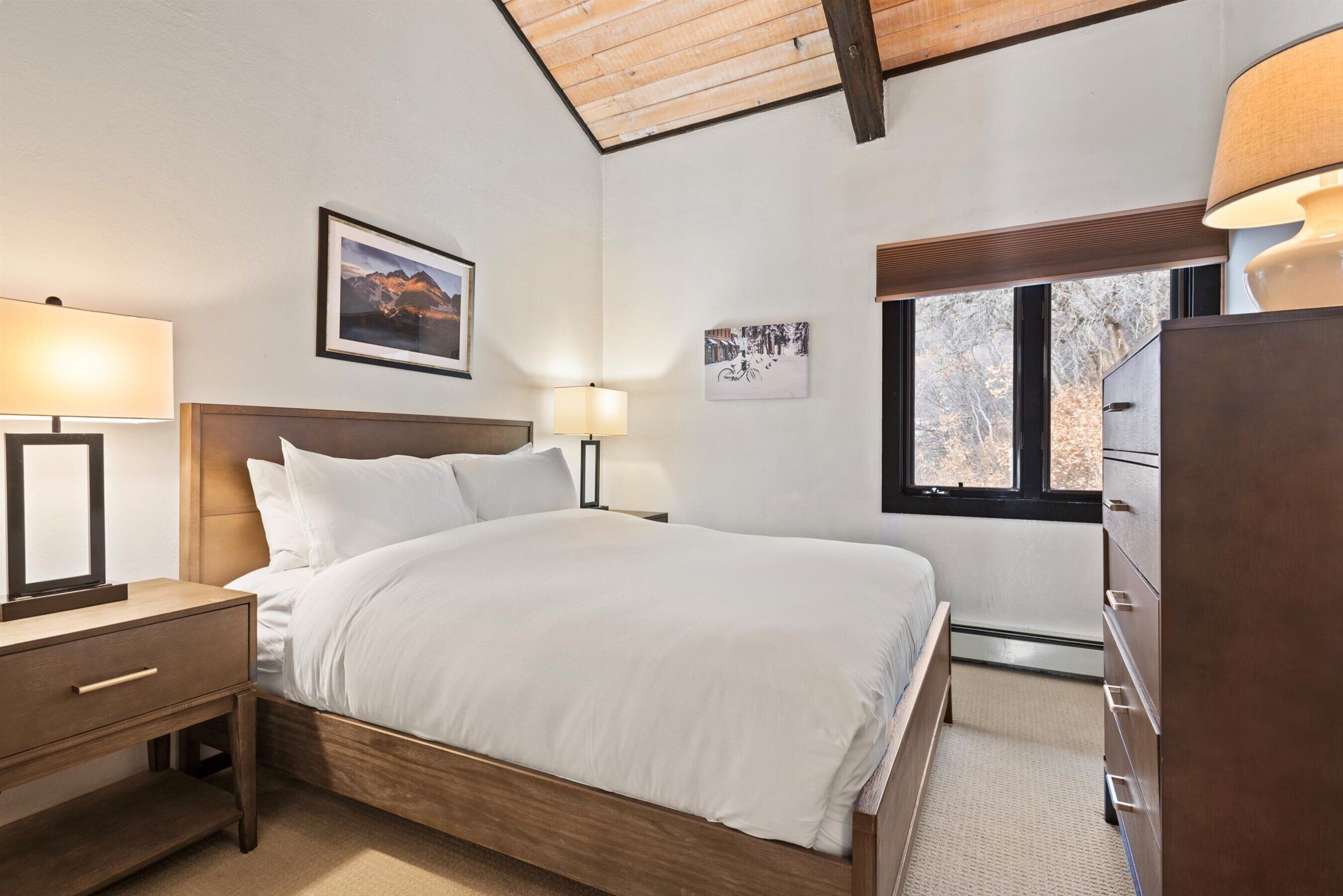 Un dormitorio con una cama de madera, dos lámparas de noche, una mesita de noche, un aparador y una ventana con vista a árboles.