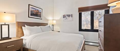 Un dormitorio con una cama de madera, dos lámparas de noche, una mesita de noche, un aparador y una ventana con vista a árboles.