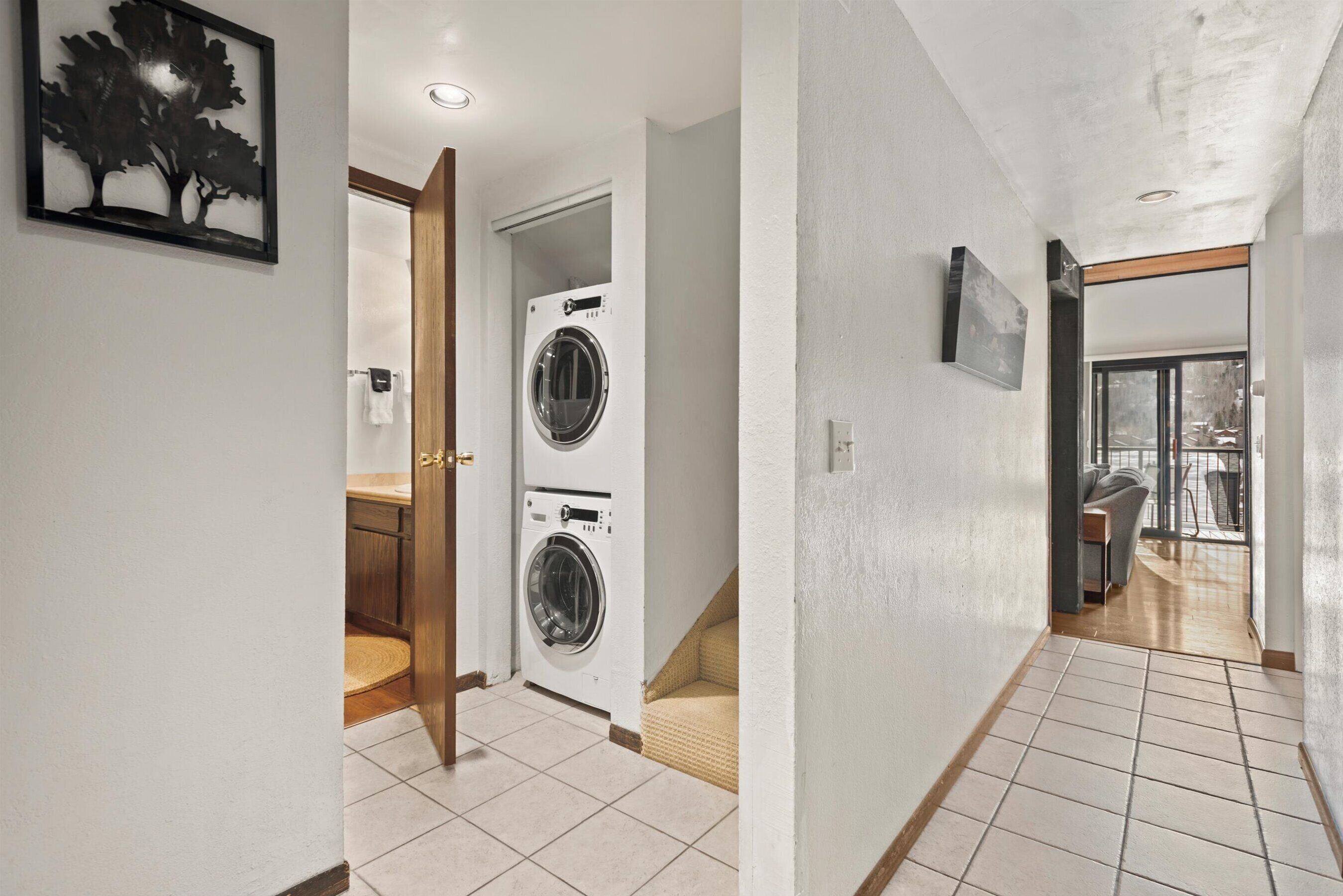 A hallway with a washing machine, a door leading to a bathroom, and a view into a living area.