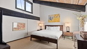 Une chambre à coucher avec un grand lit, deux tables de chevet avec des lampes, un plafond en bois et des œuvres d’art accrochées aux murs.