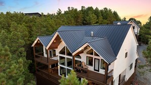 A modern cabin with a metal roof, multiple decks, and large windows, surrounded by dense pine trees. - Pool Table, Arcades, Great Views, Pets, Lazy Bear Retreat (Broken Bow)