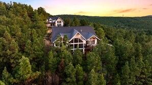 A large, two-story house surrounded by dense forest, with a prominent balcony and a second house visible in the background. - Pool Table, Arcades, Great Views, Pets, Lazy Bear Retreat (Broken Bow)
