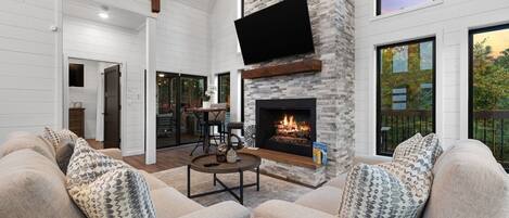 A spacious living room with a stone fireplace, a flat-screen TV mounted above it, and a large window offering a view of the outdoors.
