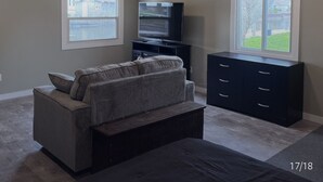 A room with a wooden ceiling, a gray couch, a TV on a stand, and a window with a view of a house and tree. - WATER FRONT & BACK 4 SEASON GETAWAY AT THE BAY.
Relax with family & friends. (Blenheim)