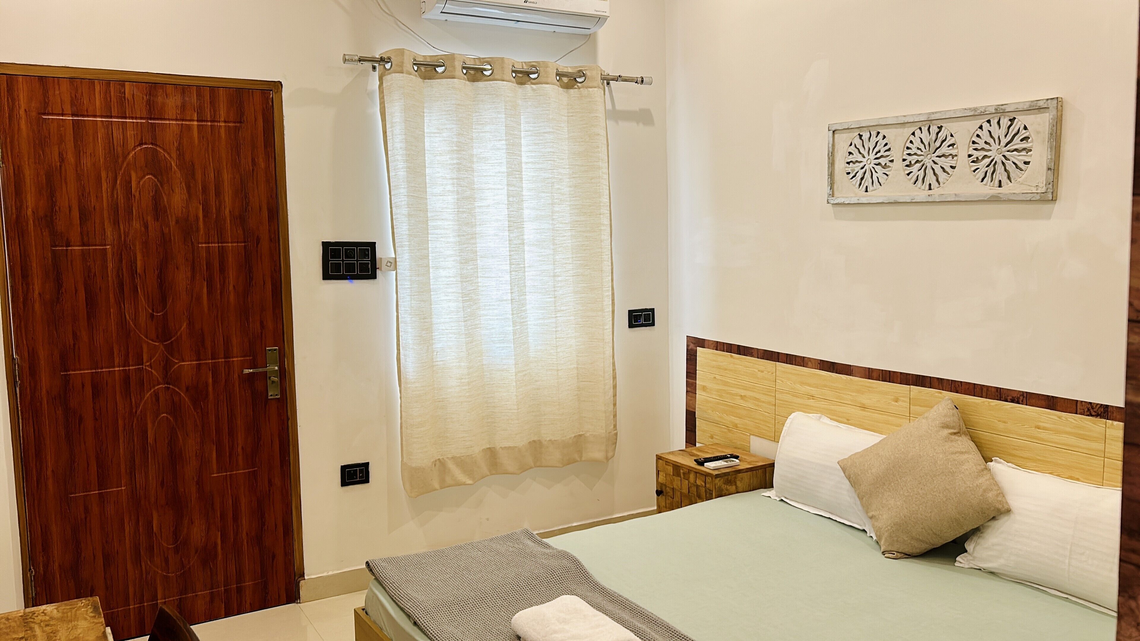 A bedroom with a wooden door, a bed with pillows, a bedside table, and a wall-mounted air conditioner.