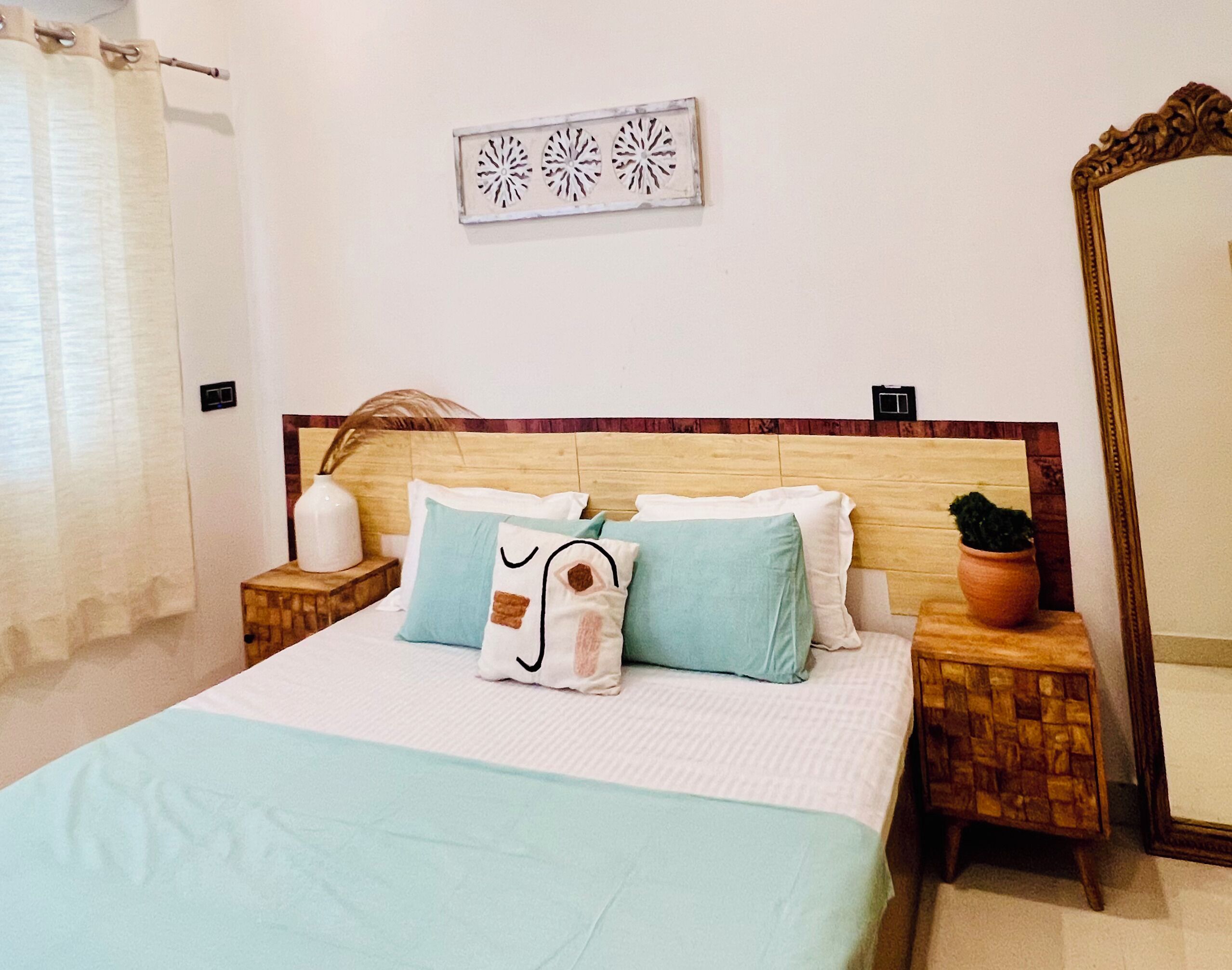 Una cama bien hecha con colcha azul claro y varios almohadones, cabecera de madera, un adorno en la pared sobre la cama, un espejo grande a la derecha y una pequeña planta en maceta en la mesita de noche.