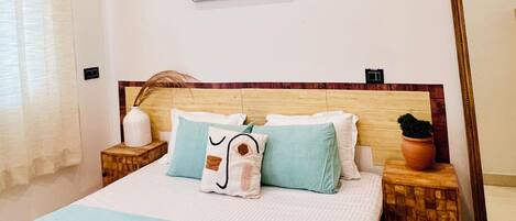 Una cama bien hecha con colcha azul claro y varios almohadones, cabecera de madera, un adorno en la pared sobre la cama, un espejo grande a la derecha y una pequeña planta en maceta en la mesita de noche.