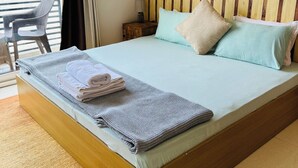 A neatly made bed with pillows and a folded towel, a wooden headboard, a small table with a lamp, and a view of the outdoors through a window. - Prithvi Hotel (Rishikesh)