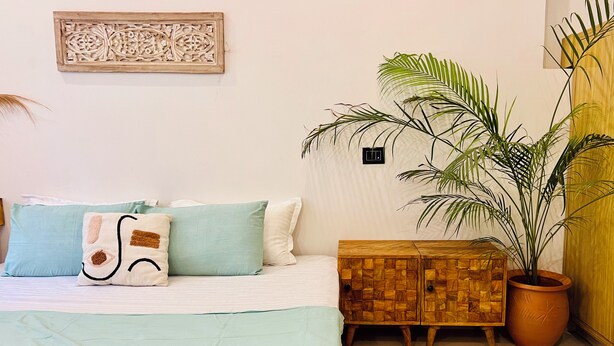 A bedroom with a bed, pillows, a wooden cabinet, a potted plant, and a wall-mounted light switch. - Prithvi Hotel (Rishikesh)
