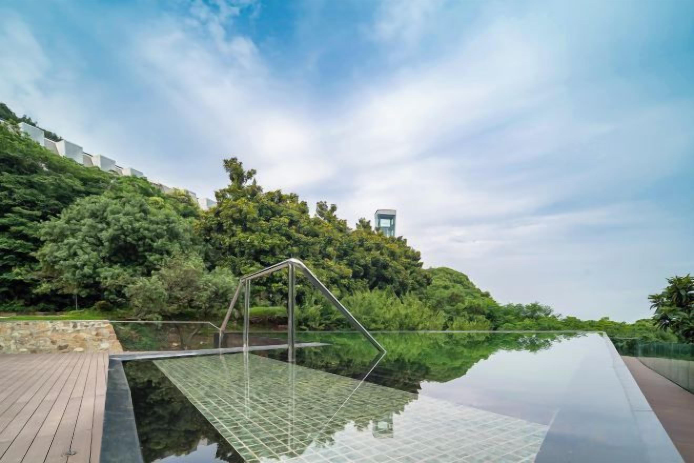 Una piscina de borde infinito de cristal con estructura metálica, rodeada por una terraza de madera y exuberante vegetación.