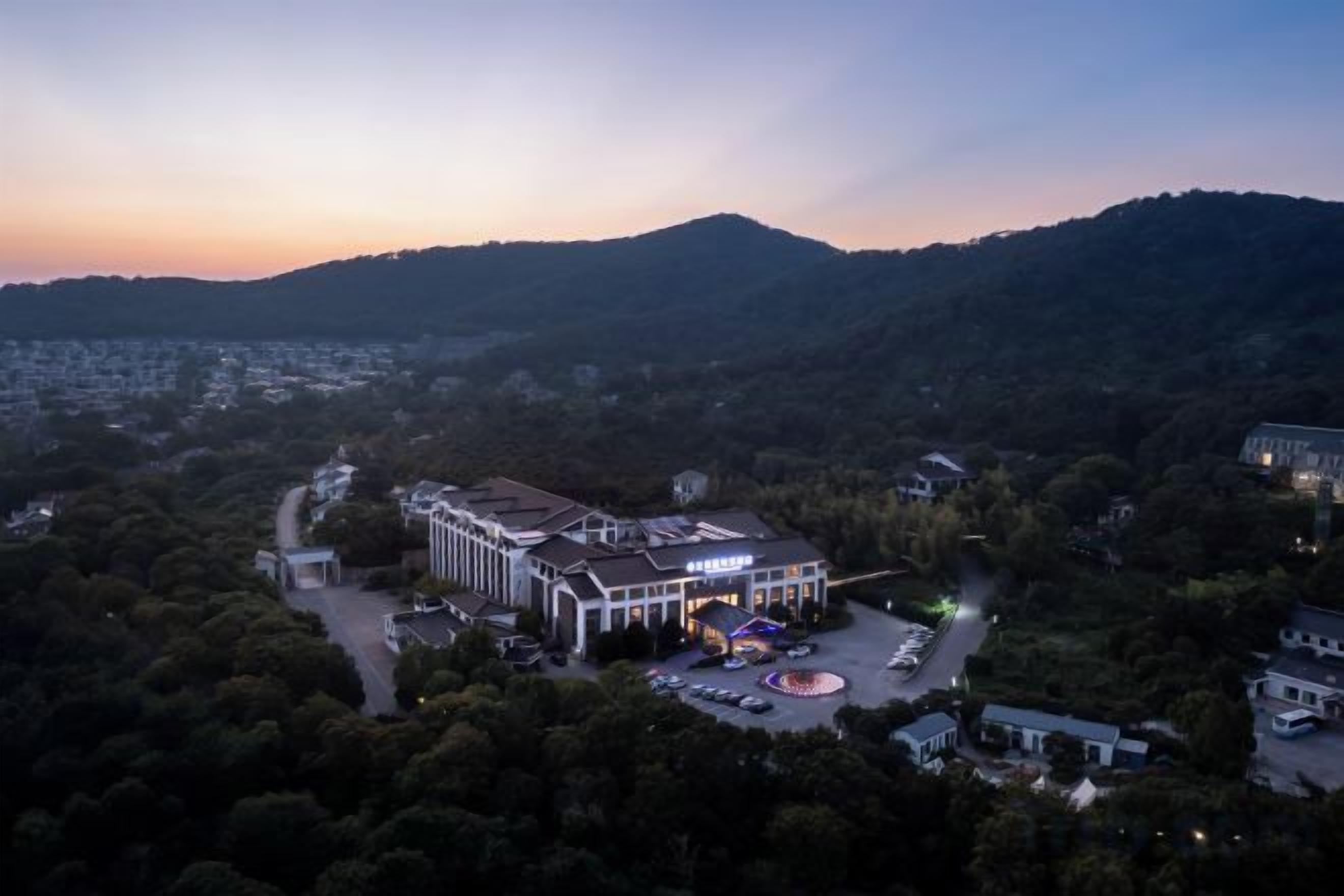 A hotel with a central building surrounded by parking and a landscaped area, set against a backdrop of hills and trees.