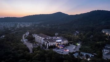 A hotel with a central building surrounded by parking and a landscaped area, set against a backdrop of hills and trees.