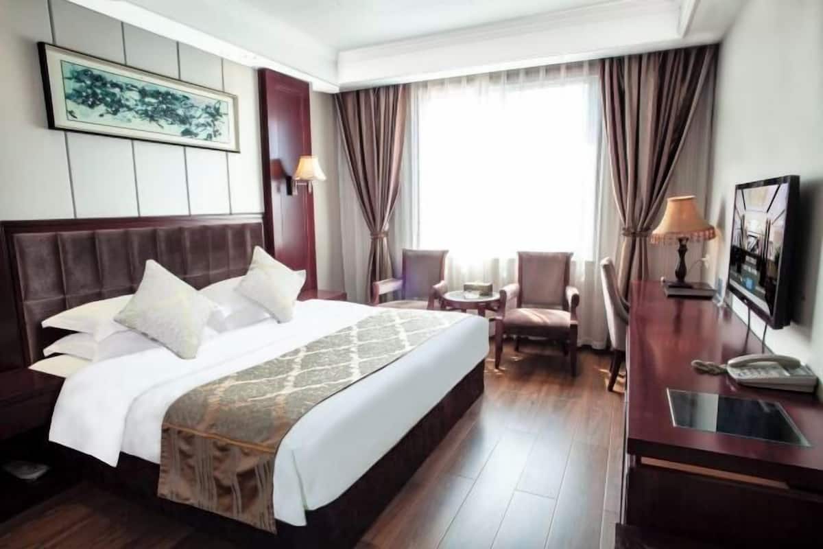 A hotel room with a large bed, a desk, a chair, a small table, and a window with curtains.