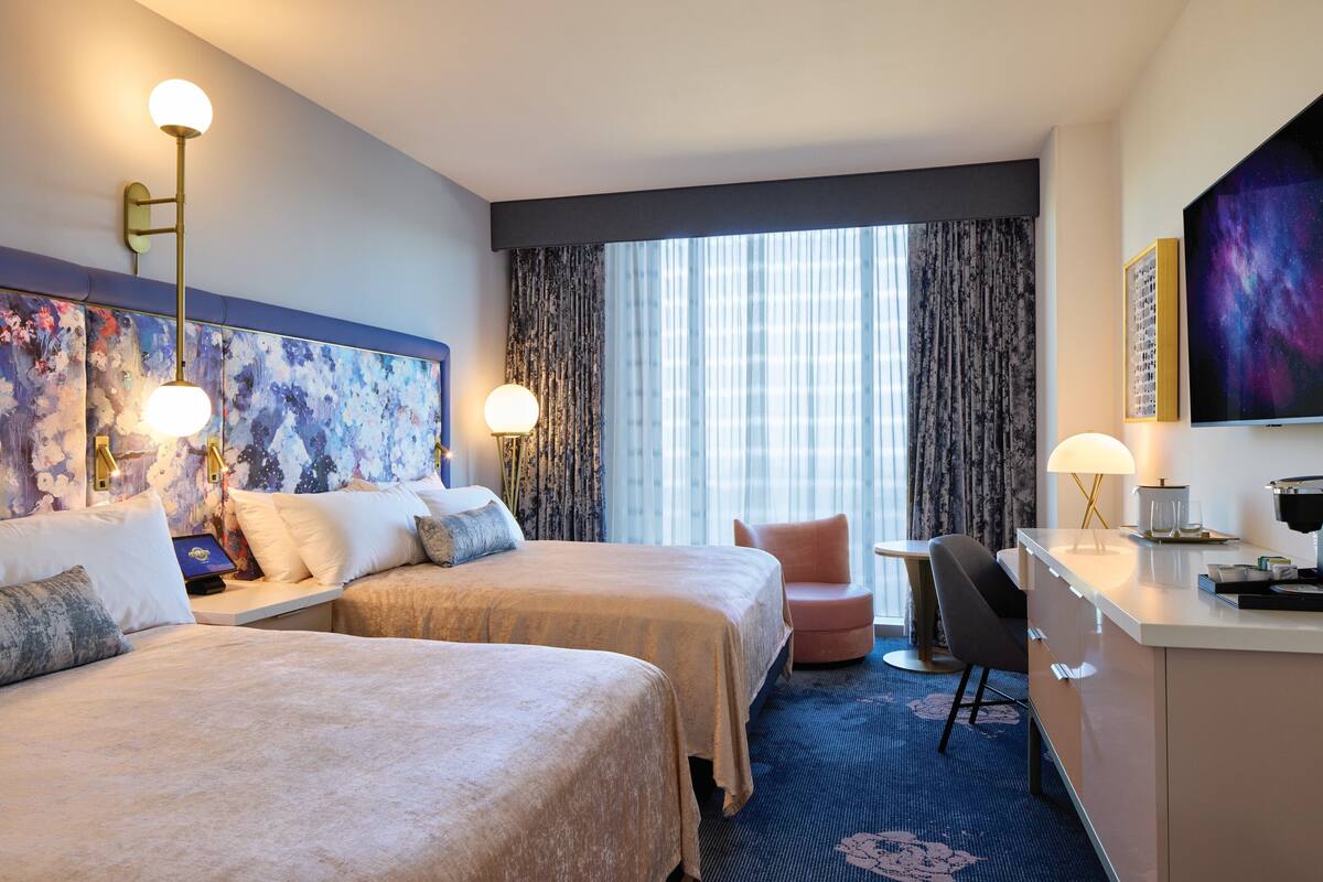 A hotel room with two beds, a desk, a chair, a large window with curtains, and artwork on the walls.