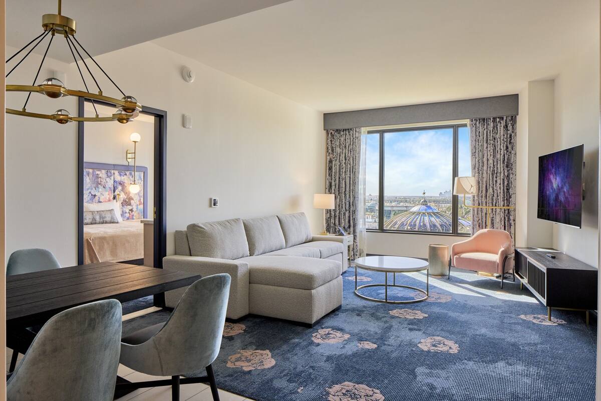 A modern hotel room with a sofa, a dining table, a TV, and a large window with a view.
