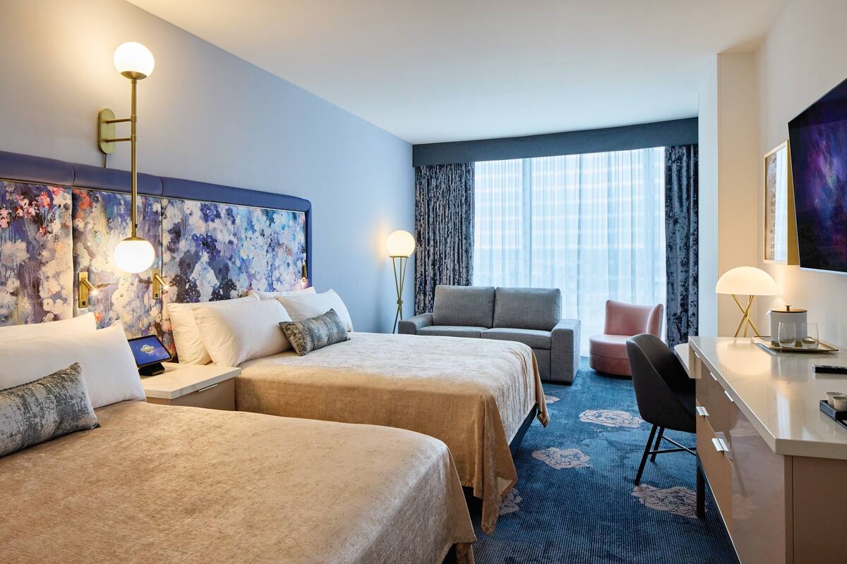A hotel room with two beds, a desk, a chair, a sofa, and a large window with curtains.