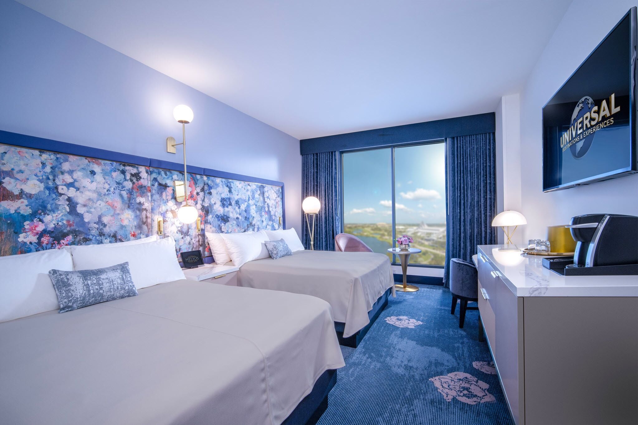 A hotel room with two beds, a desk, a chair, a large window with a view, and a wall-mounted TV displaying 'Universal'.