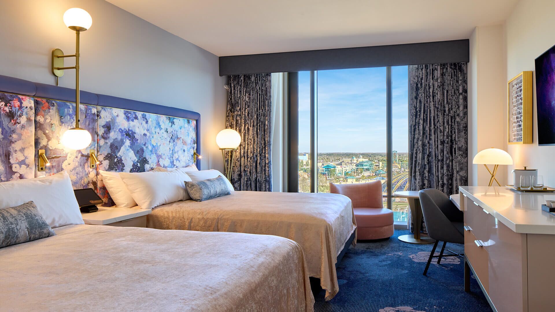 A hotel room with two beds, a desk, and a chair. There is a large window with a city view.