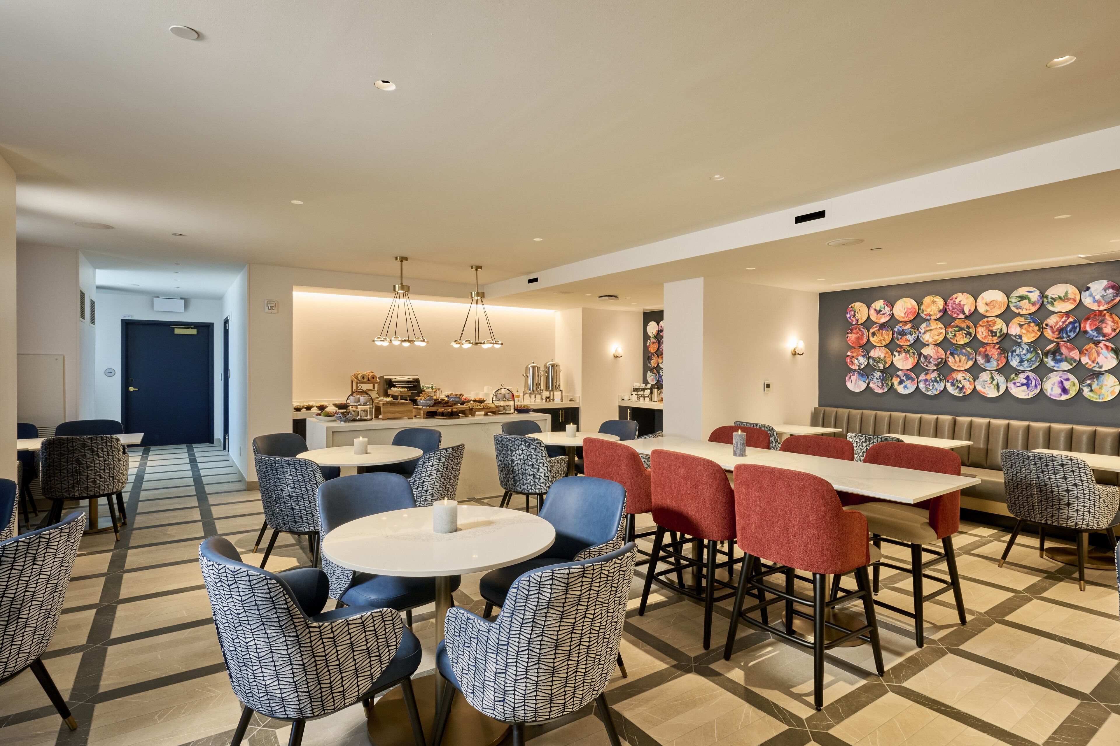 Zona pranzo moderna con tavoli e sedie rotonde, una cucina sullo sfondo e una parete decorata con piatti.