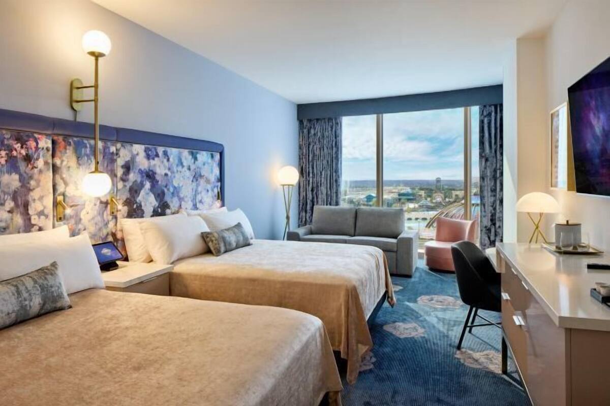 A hotel room with two beds, a sofa, a desk, and a large window offering a city view.