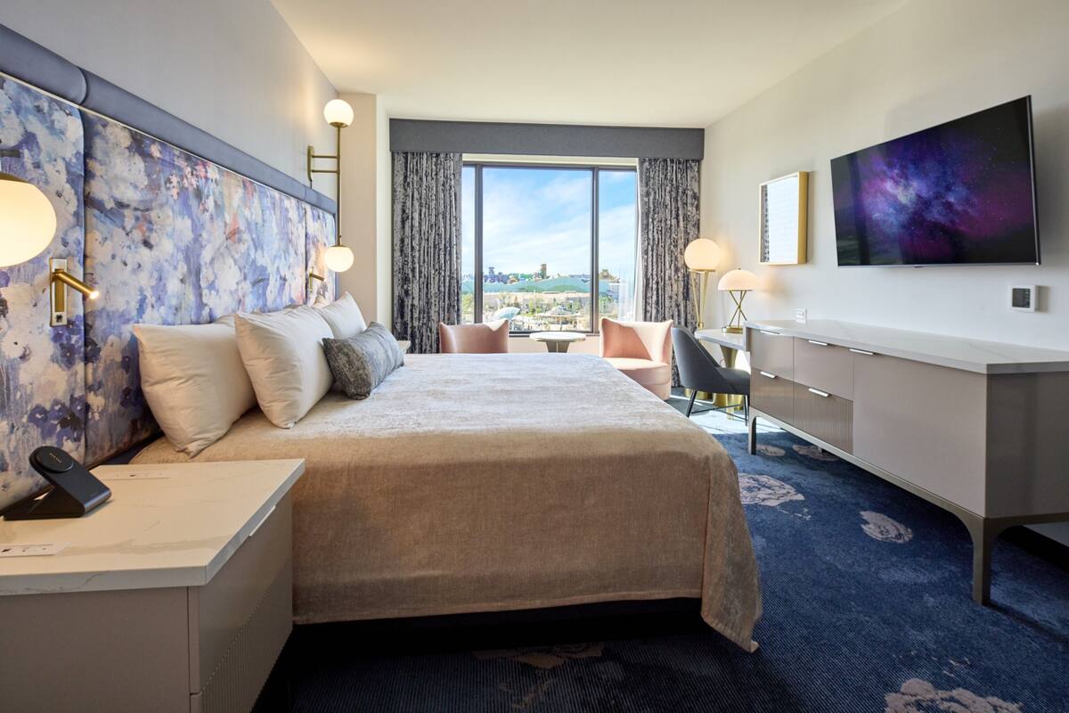 A hotel room with a large bed, a desk with a phone, a TV, and a view of the city.
