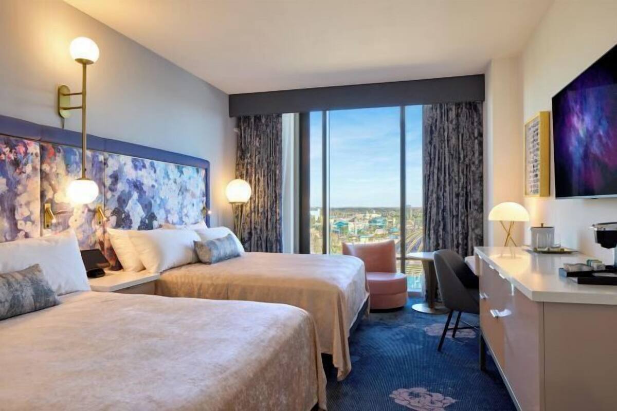 A hotel room with two beds, a desk, a chair, and a large window offering a city view.