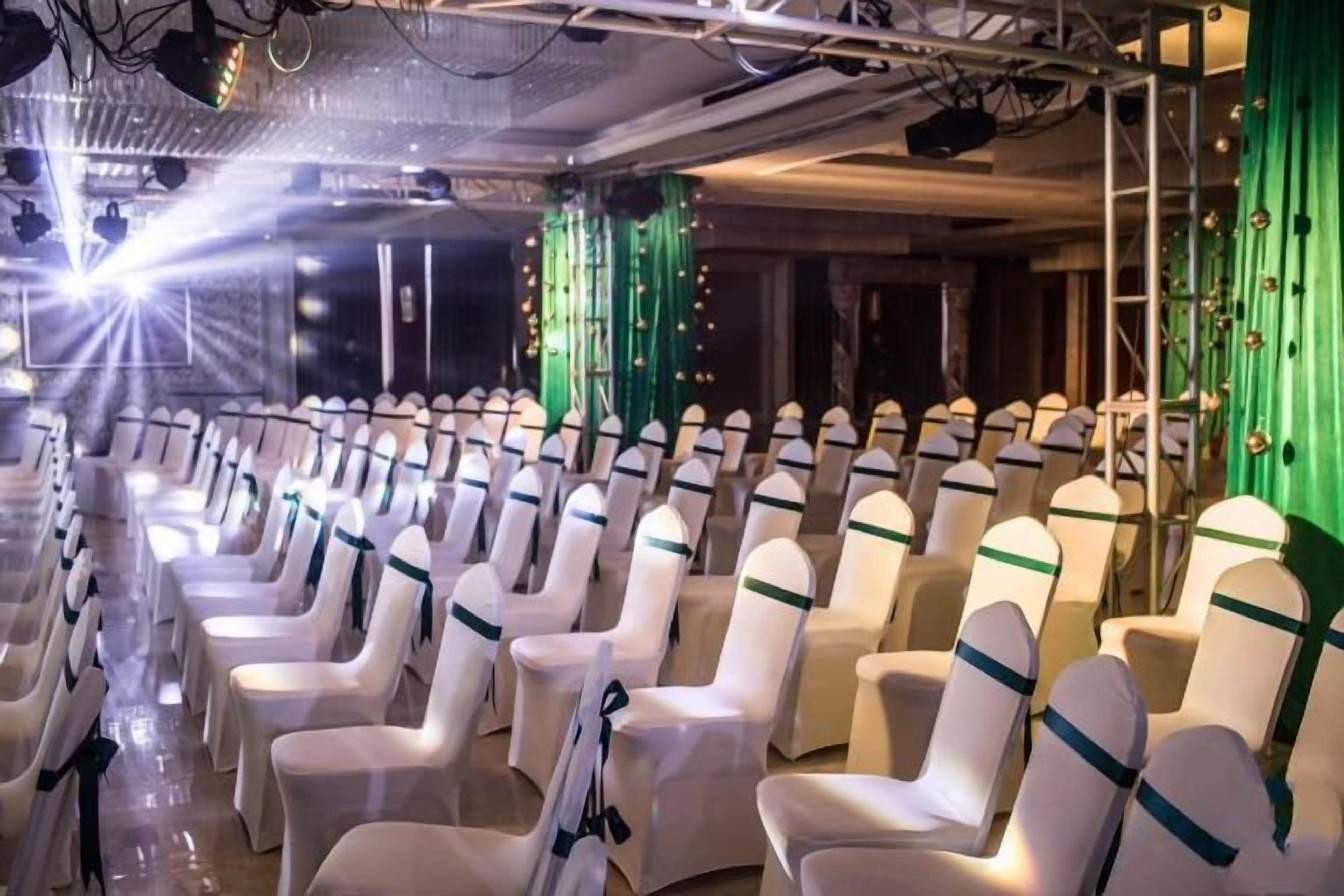 Filas de sillas con fundas blancas y cintas verdes, dispuestas en un salón de banquetes.