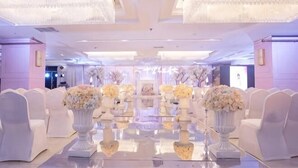 A formal event space with white chairs, reflective tables, and large floral centerpieces.