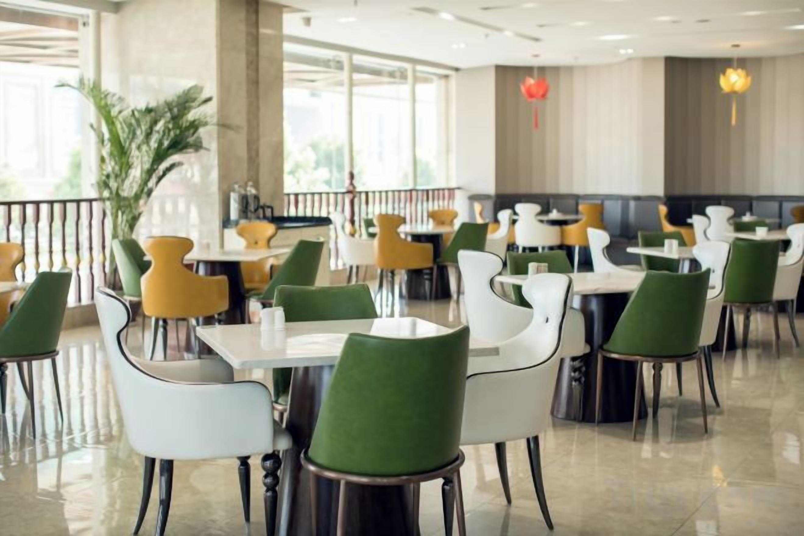 A restaurant with multiple tables and chairs, a potted plant, and a view through large windows.