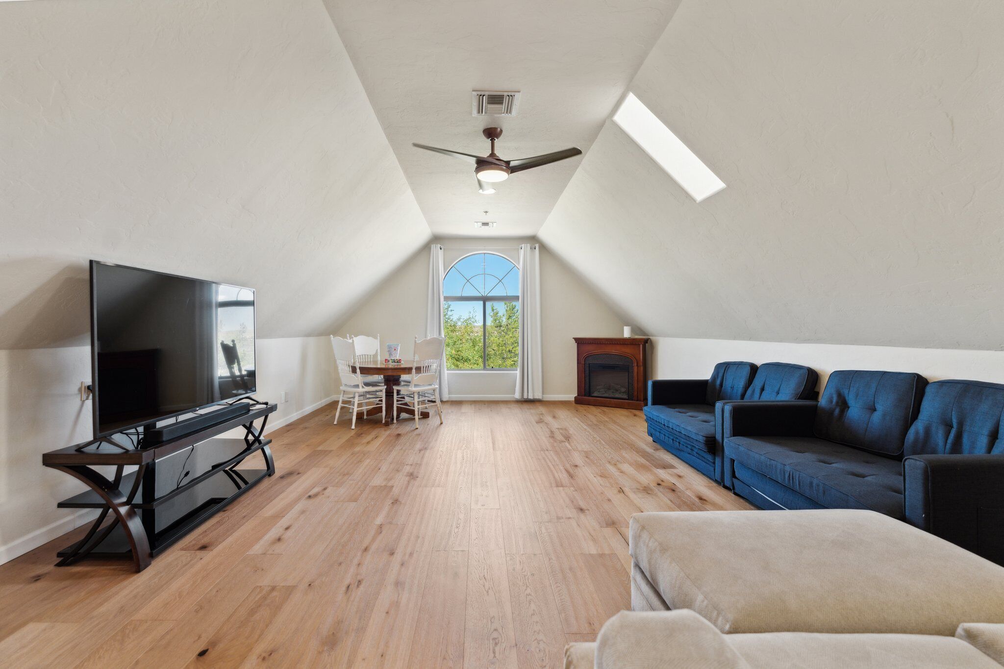 A spacious attic room with a wooden floor, a ceiling fan, a large TV, a blue sofa, and a fireplace.