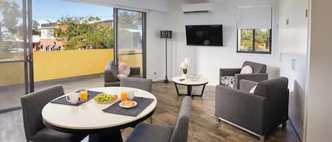A modern living room with a round dining table, grey sofas, and a flat-screen TV mounted on the wall.