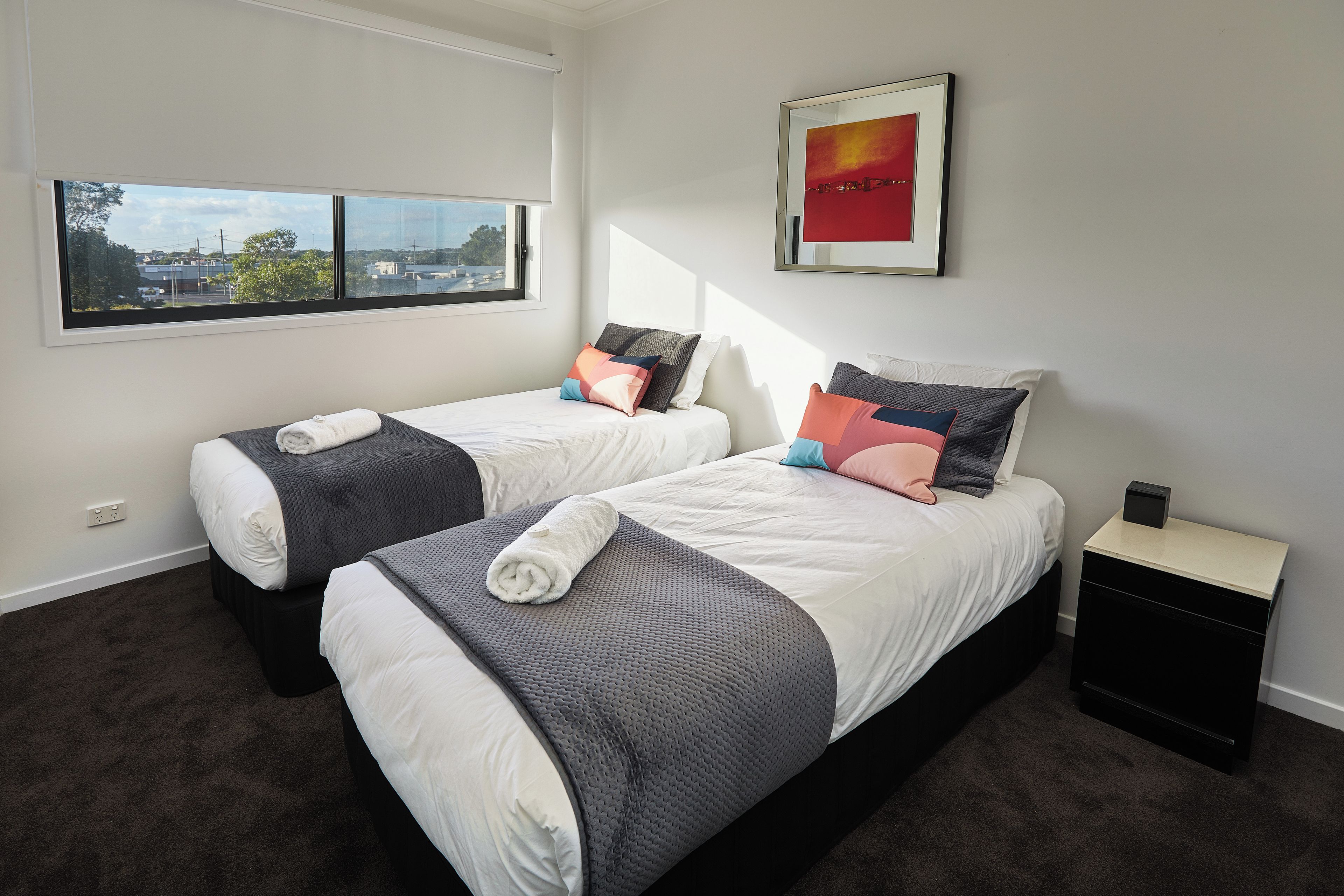A hotel room with two beds, a nightstand, a framed picture, and a window with a city view.